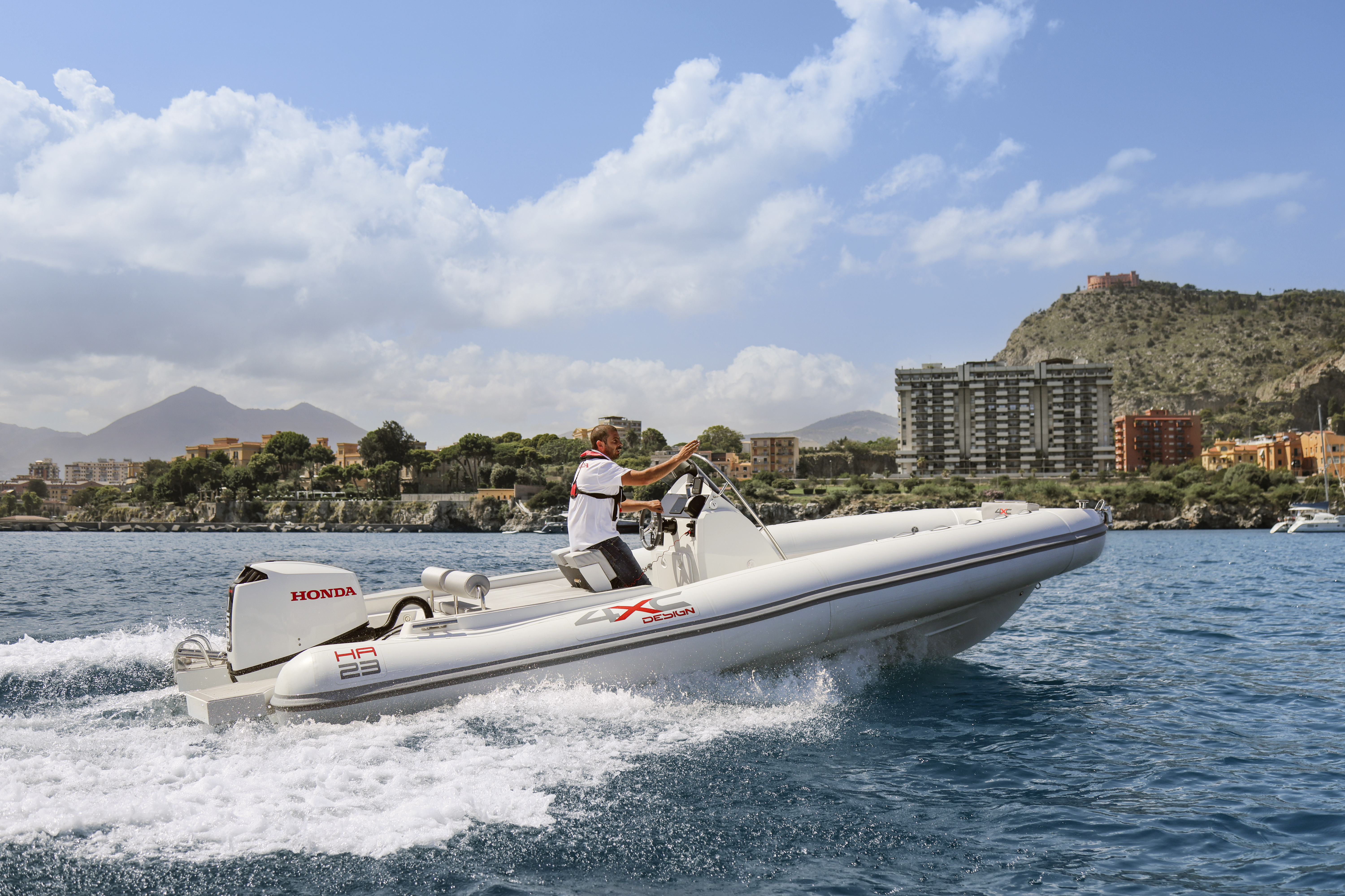 A white inflatable speedboat with a Honda outboard motor races across blue waters near a coastal city. The boat is piloted by a person in white clothing. In the background, a shoreline with buildings and mountains is visible under a partly cloudy sky.
